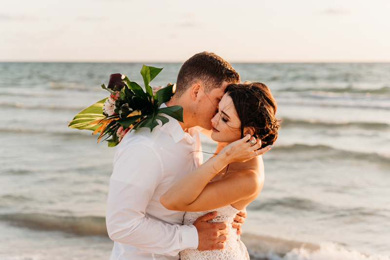 Wedding Photographer, Bride and groom embrace, she still holds bouquet, they are at beach