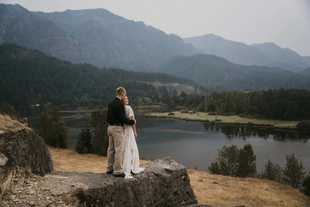 A bride and groom stands on a cliff overlooking the mountains of the columbia river gorge.