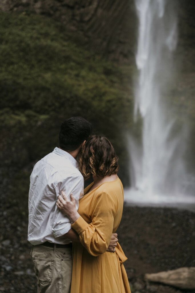 couple stands at the base of a waterfall looking out