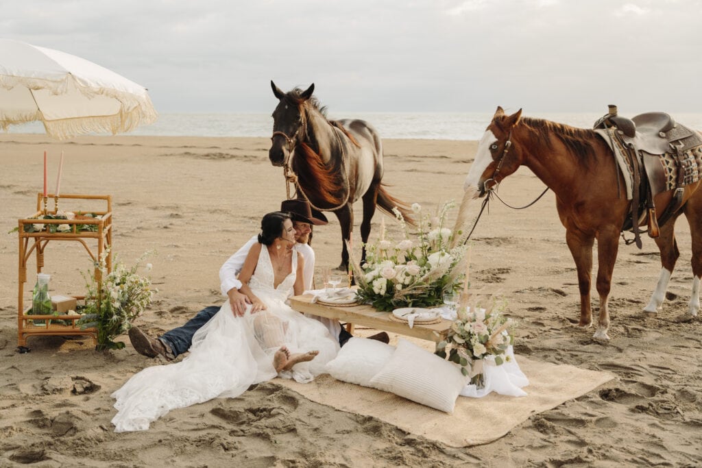 Bride and groom sit on the beach at a picnic with flowers and their two horses