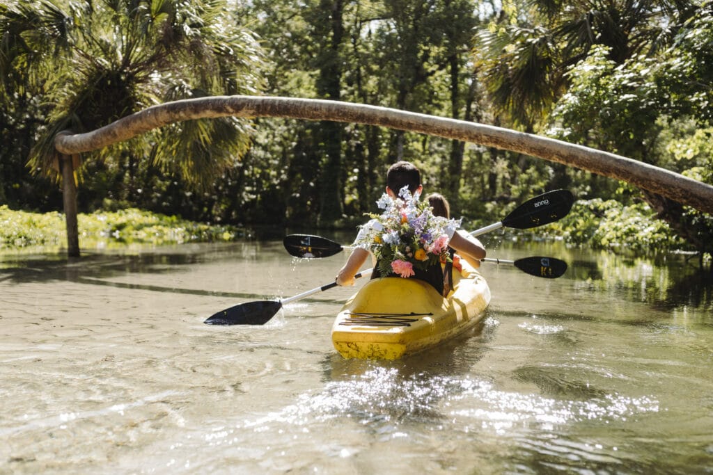 couple kayaks down clear stream with wedding bouquet in tow